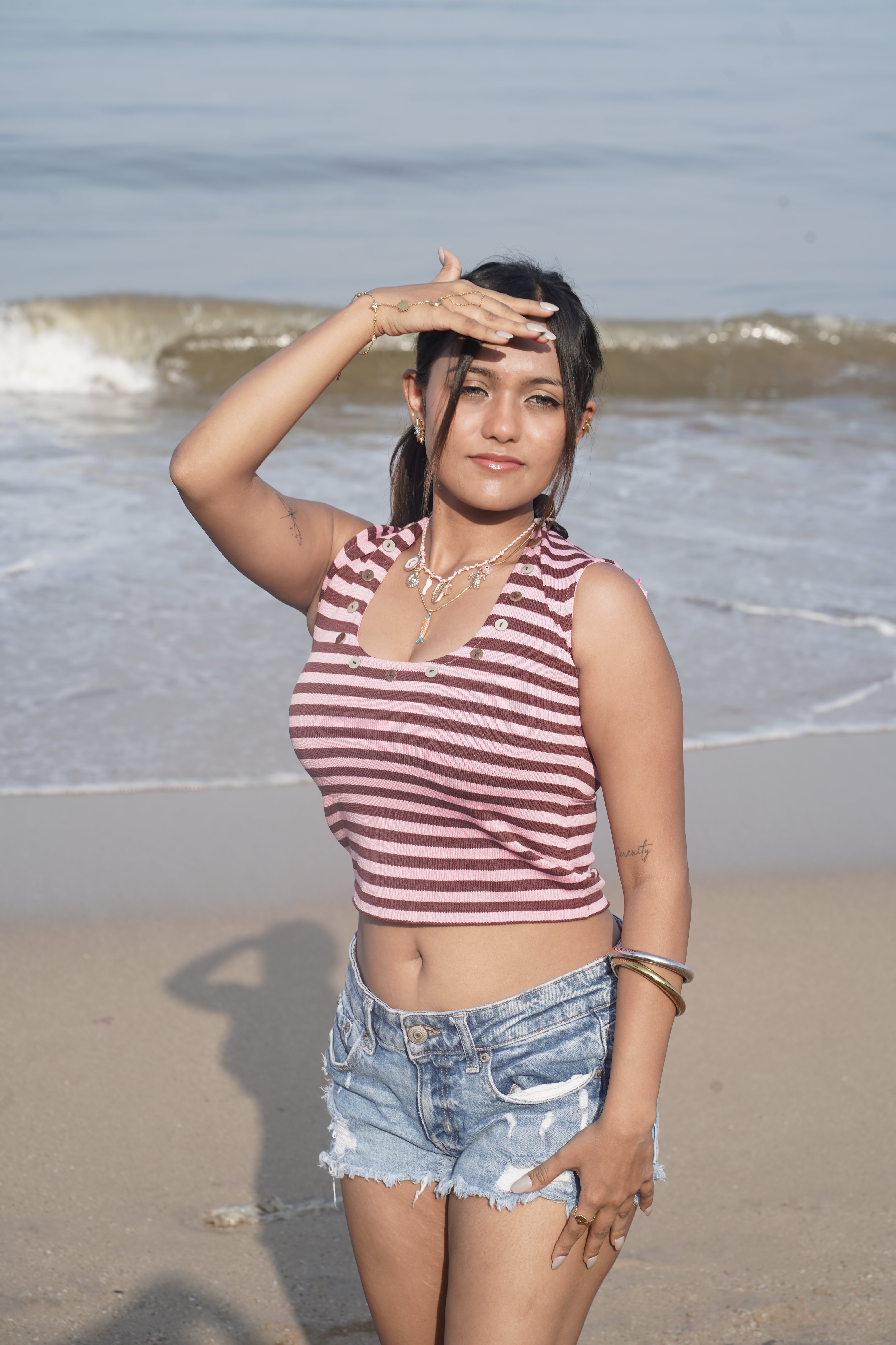 Woman in a striped tank top and denim shorts standing on a beach with ocean waves in the background.
